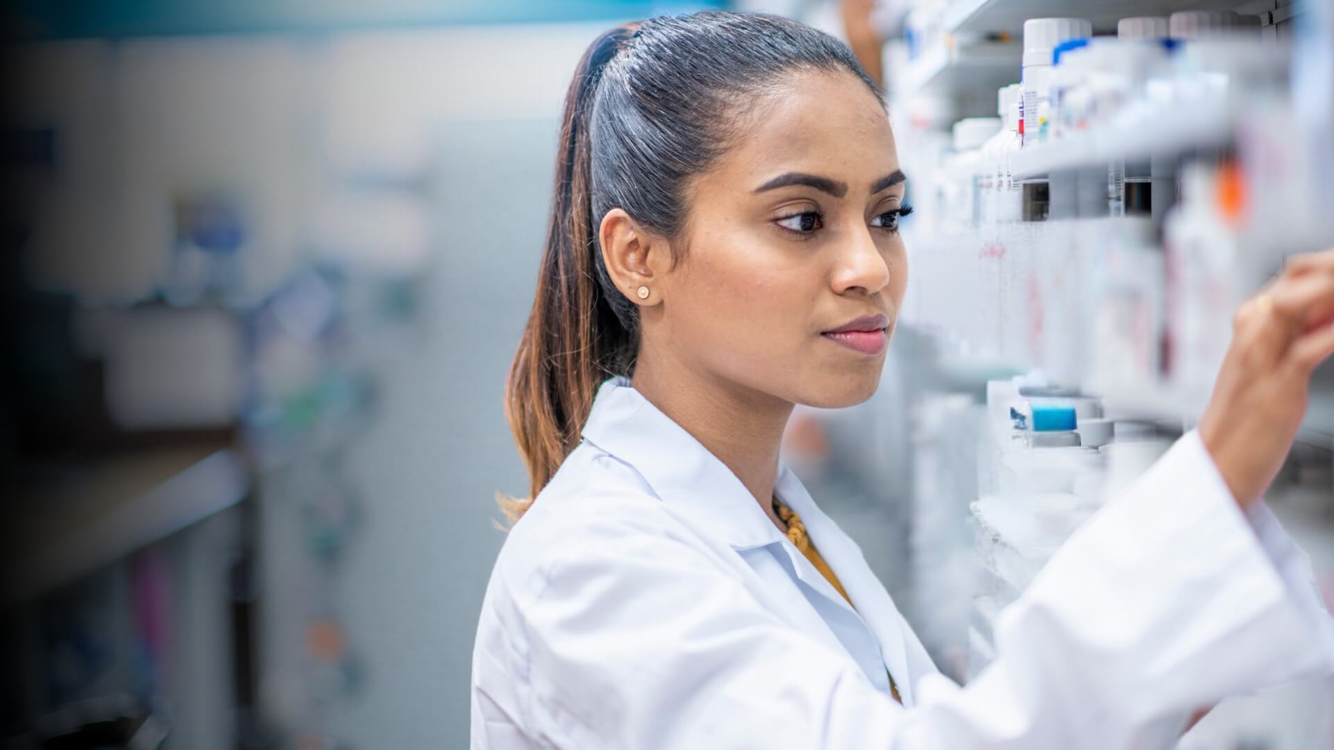 Pharmacist in a white lab coat organizing medication bottles on shelves in a pharmacy.