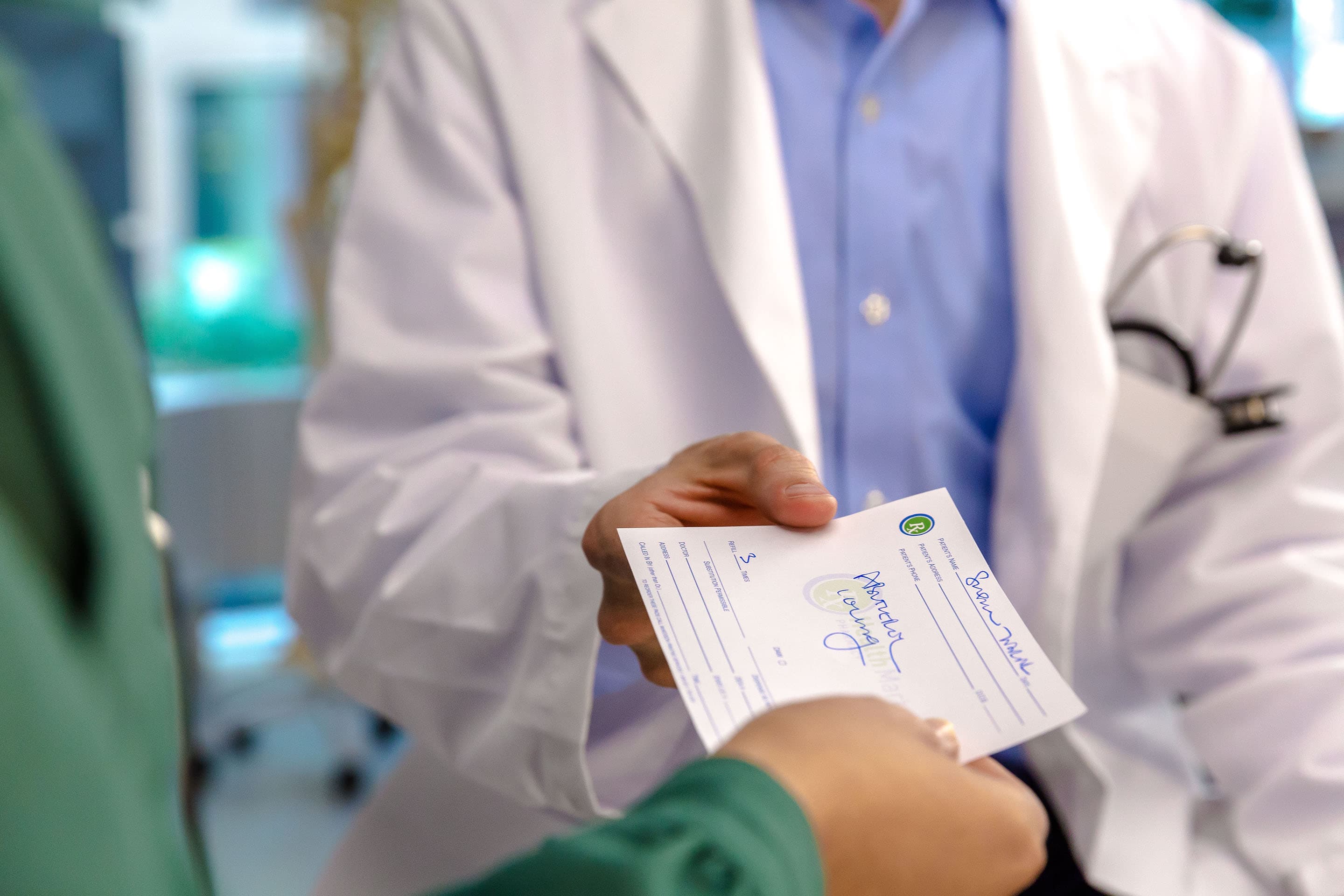 A doctor in a white coat hands a prescription to a person wearing green scrubs.