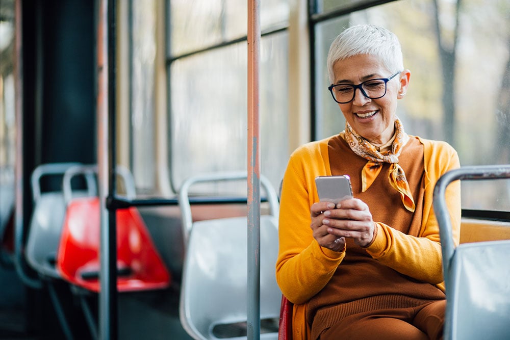 Person with short white hair, wearing a yellow sweater and scarf, sits on a bus looking at a smartphone. The bus seats are mostly empty.