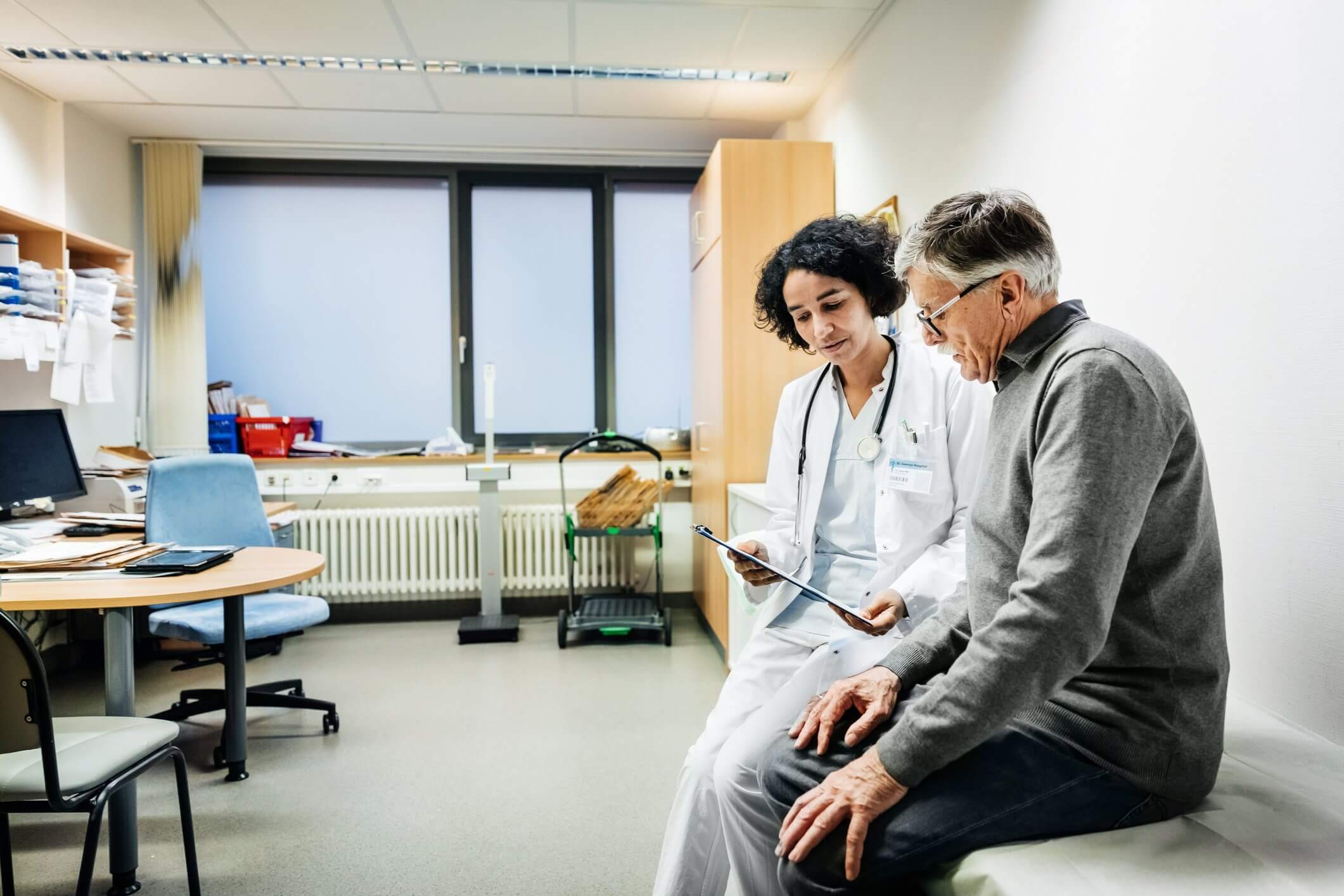 A doctor and a patient sit in a medical office discussing something on a clipboard.