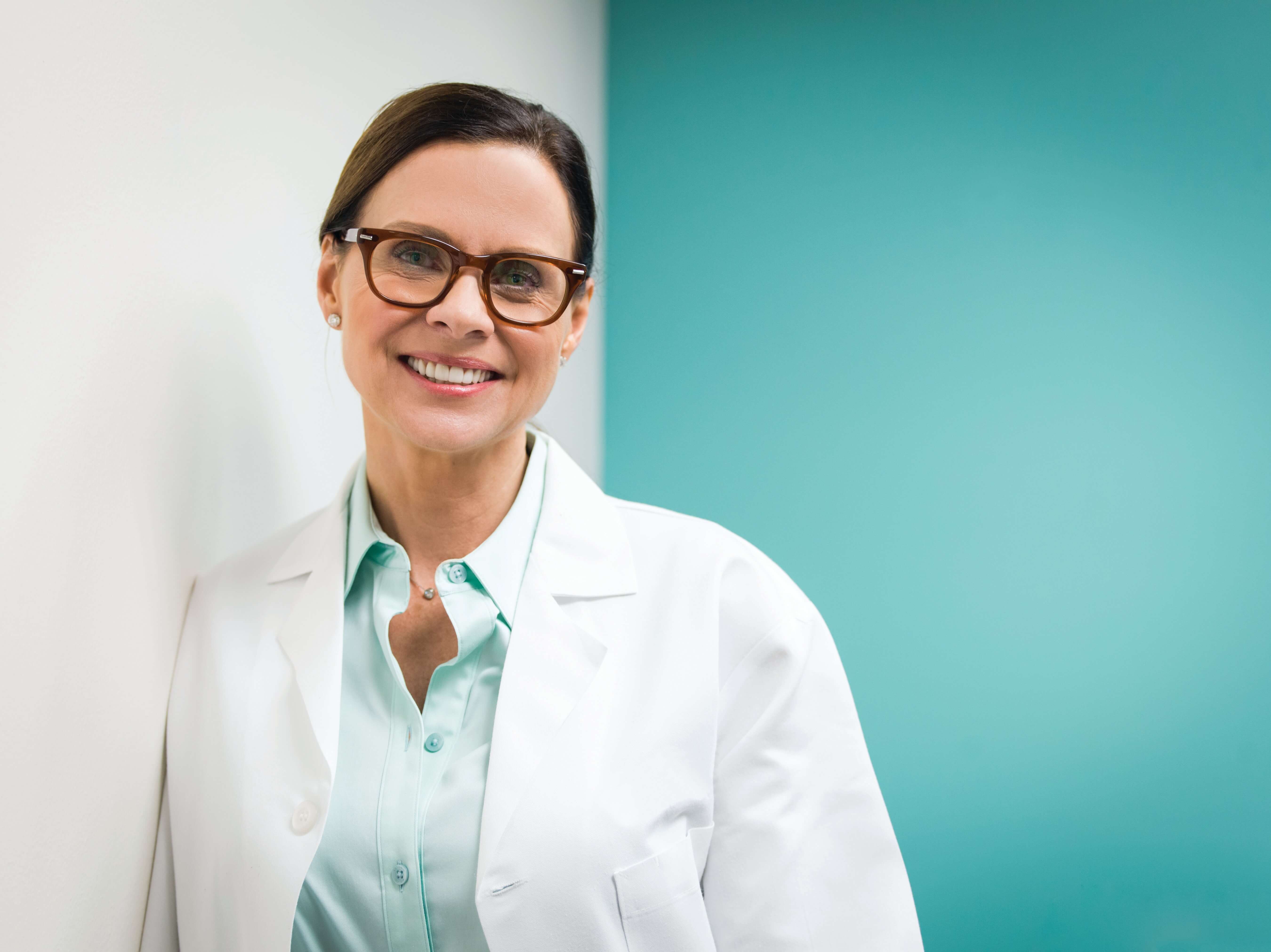 A person in a white lab coat stands against a teal wall.