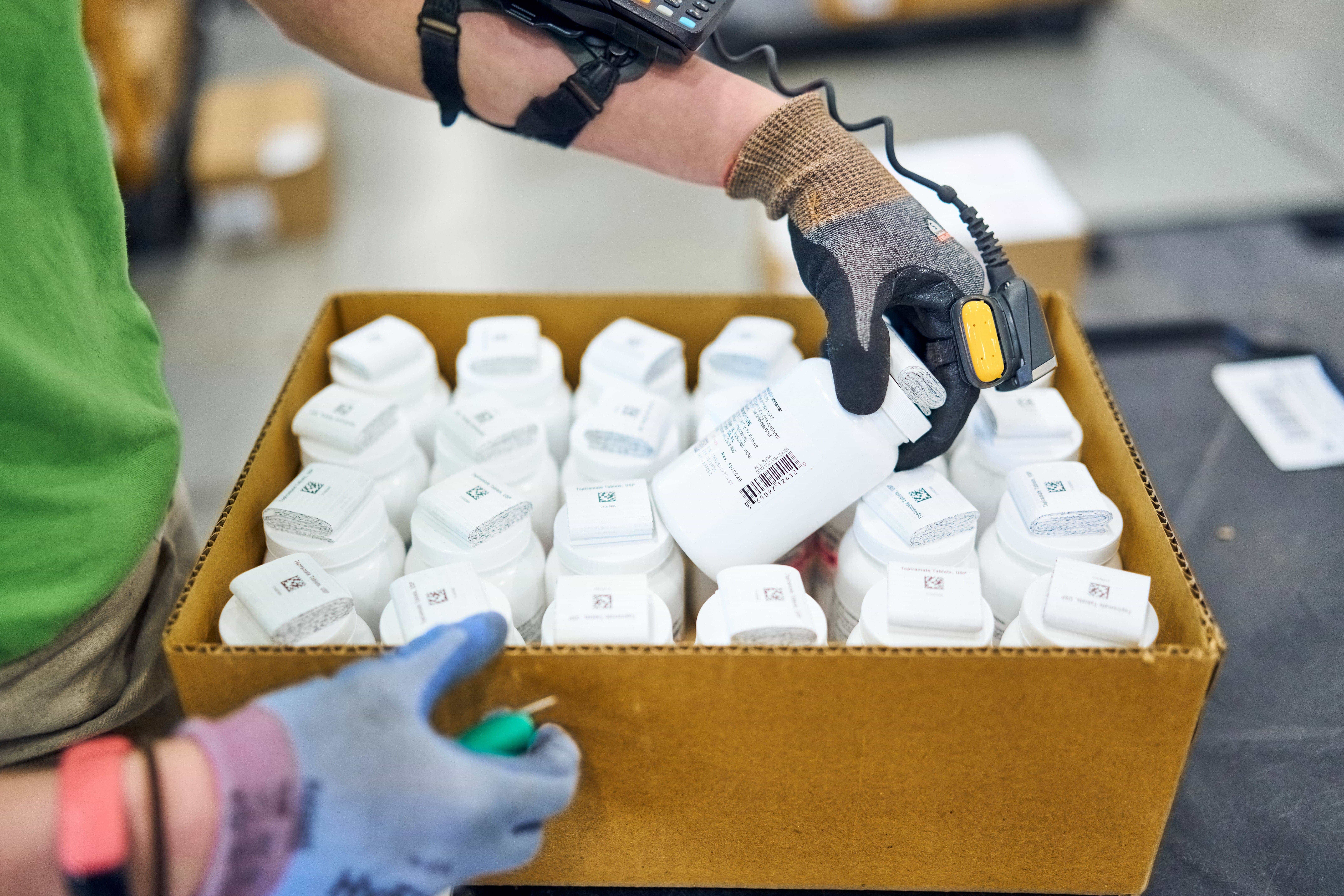 A person wearing gloves scans a bottle from a box filled with identical white bottles.