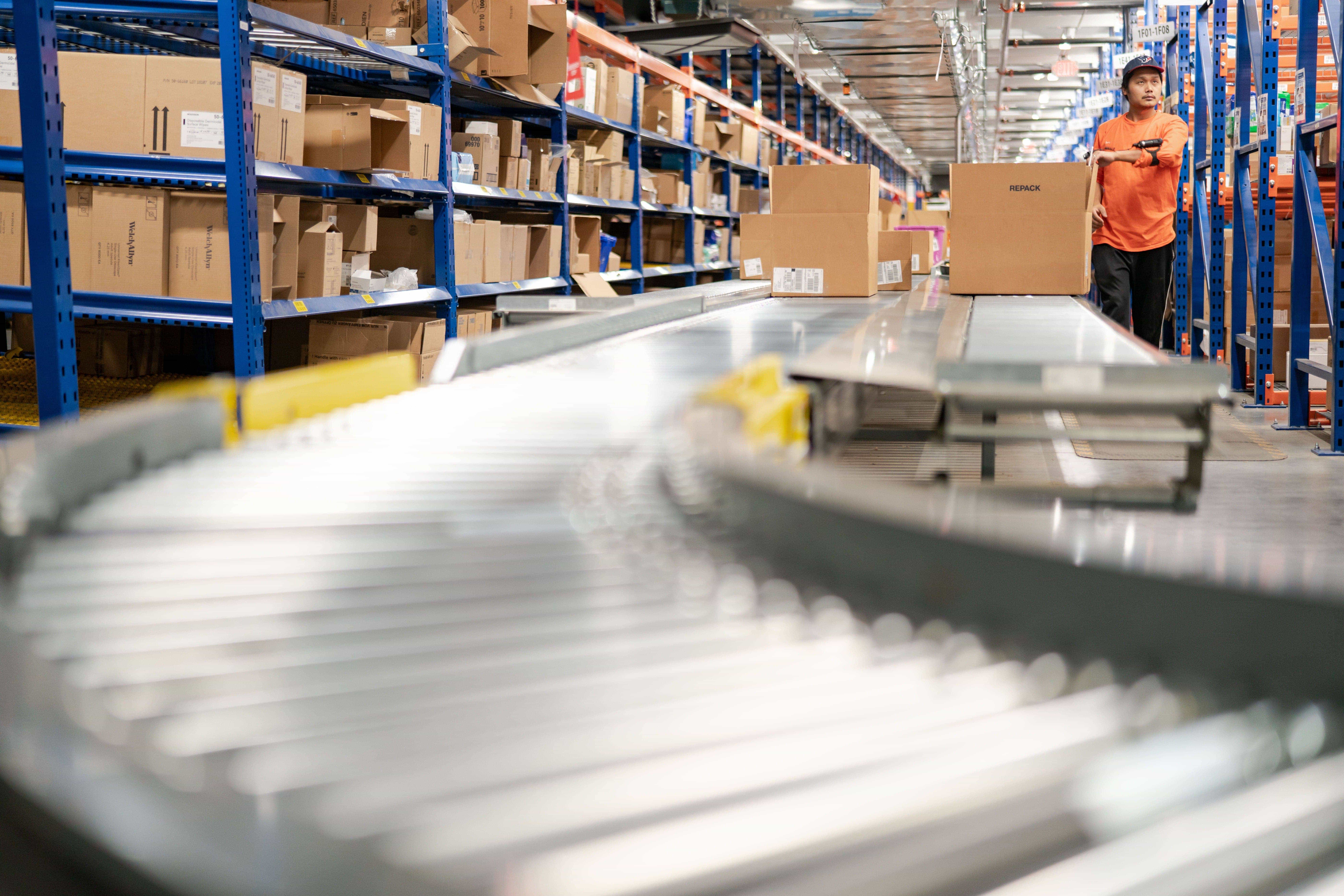 A warehouse with shelves of boxes and a conveyor belt in the foreground. A person in an orange shirt is handling a box.