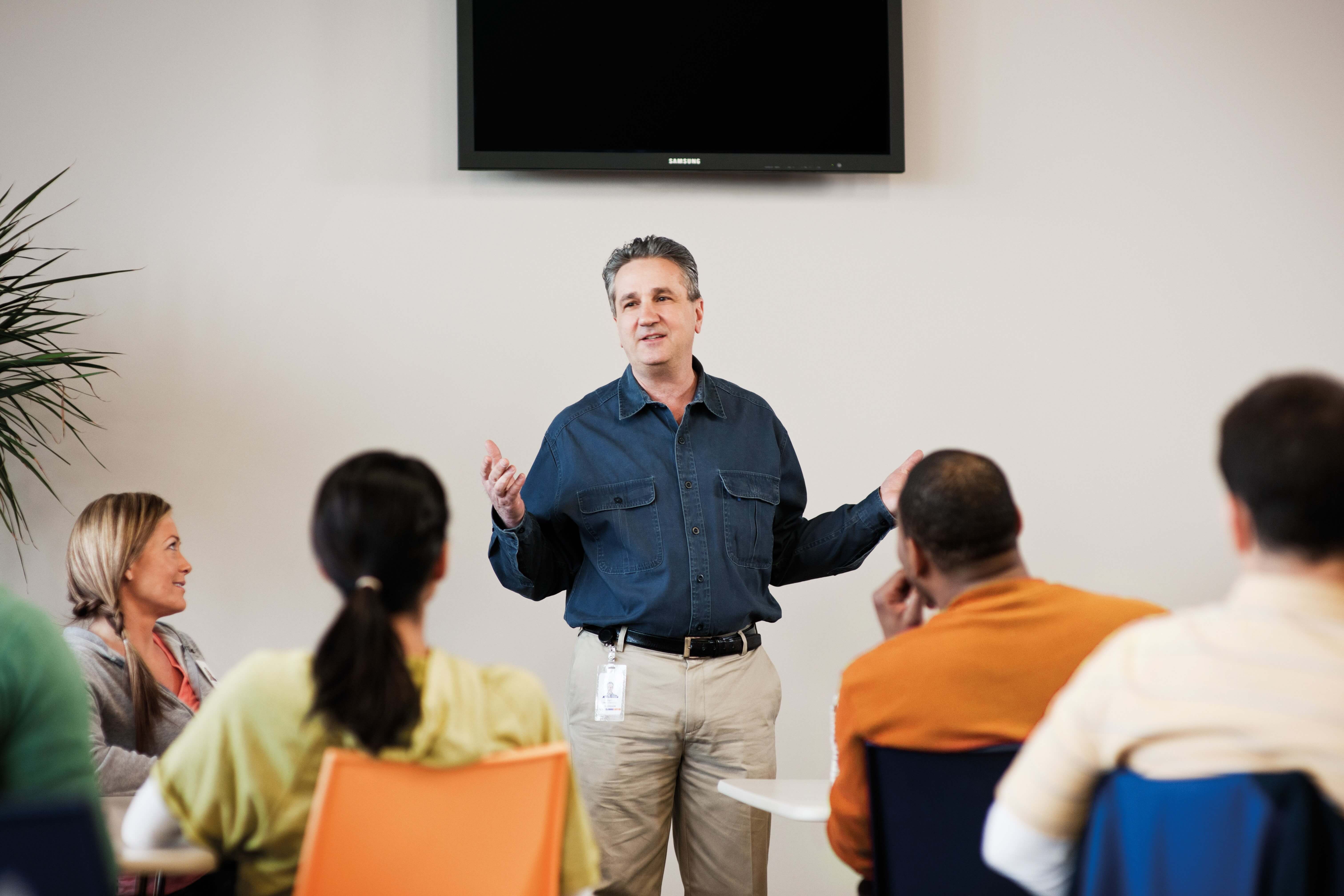 A person in a blue shirt speaks to a group of seated individuals in a classroom setting.