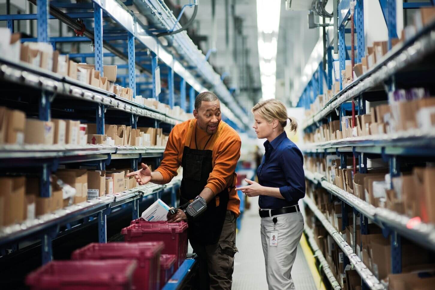 Two people in a warehouse aisle discuss inventory surrounded by shelves filled with boxes.