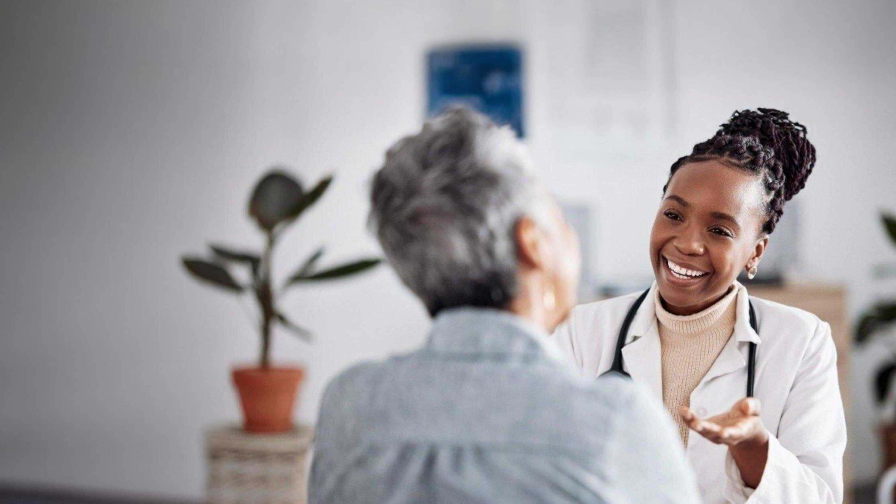 A doctor in a white coat speaks with an older person in a room with a potted plant.