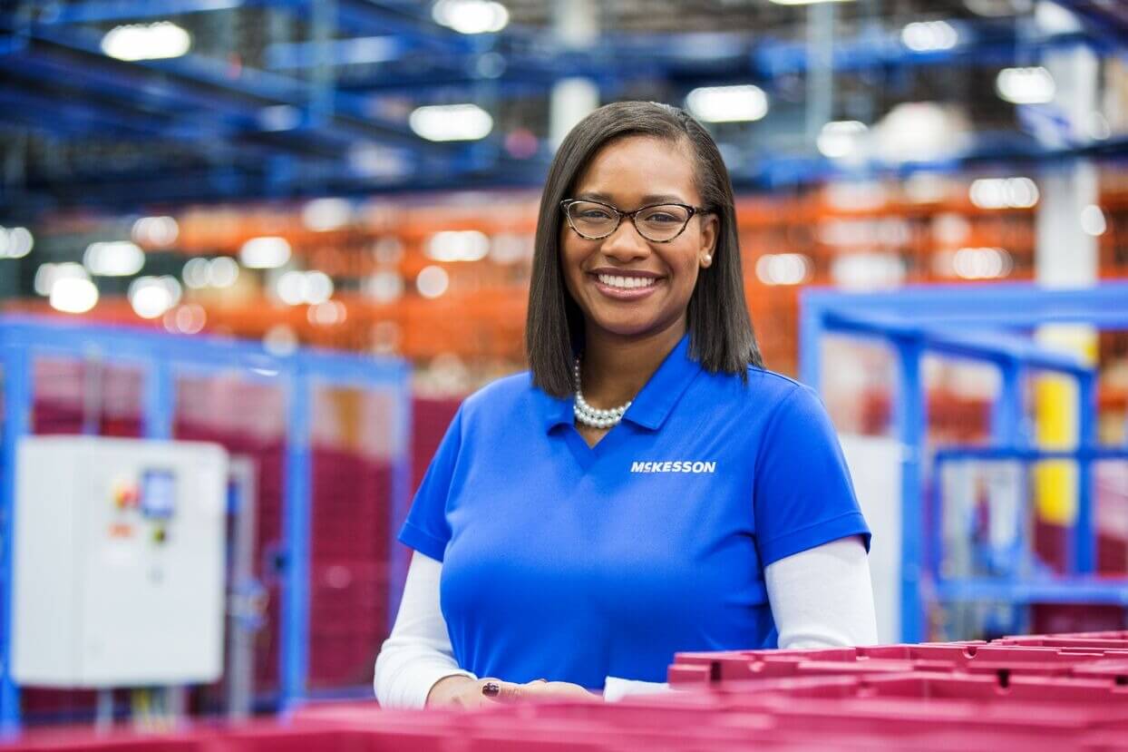 A person in a blue McKesson shirt stands in a warehouse with red bins and blue shelving.
