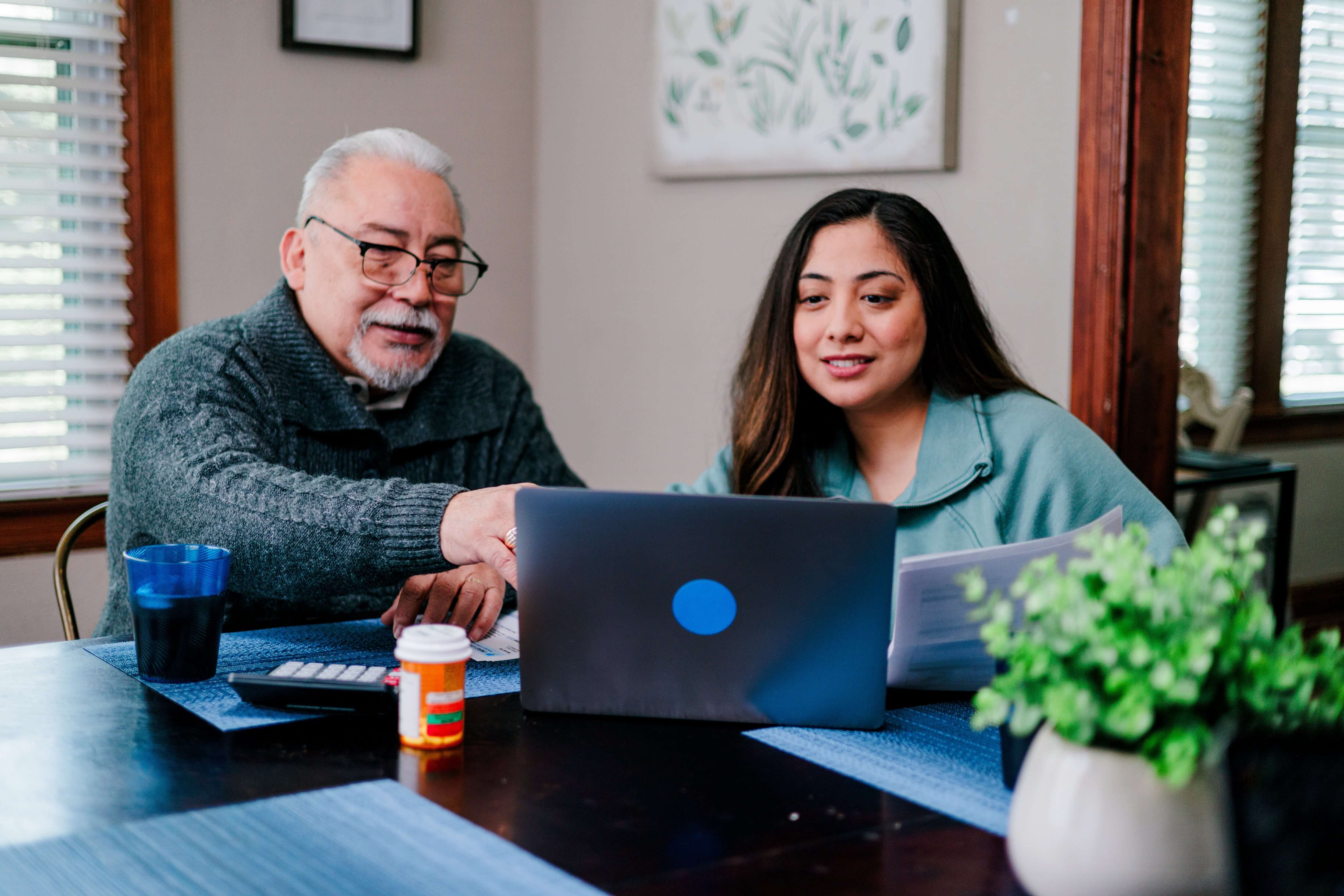 Two people sitting at a table with a laptop, documents, and a prescription bottle.