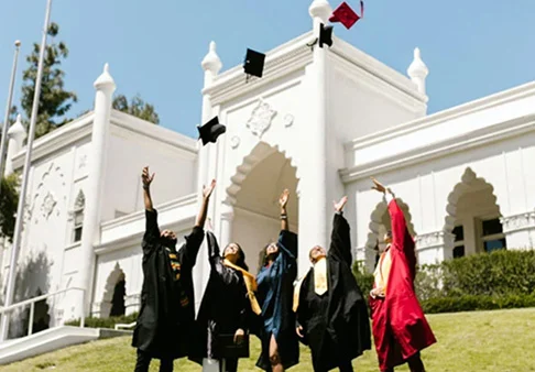 Graduates in caps and gowns celebrating by throwing their caps in the air outside a white building.