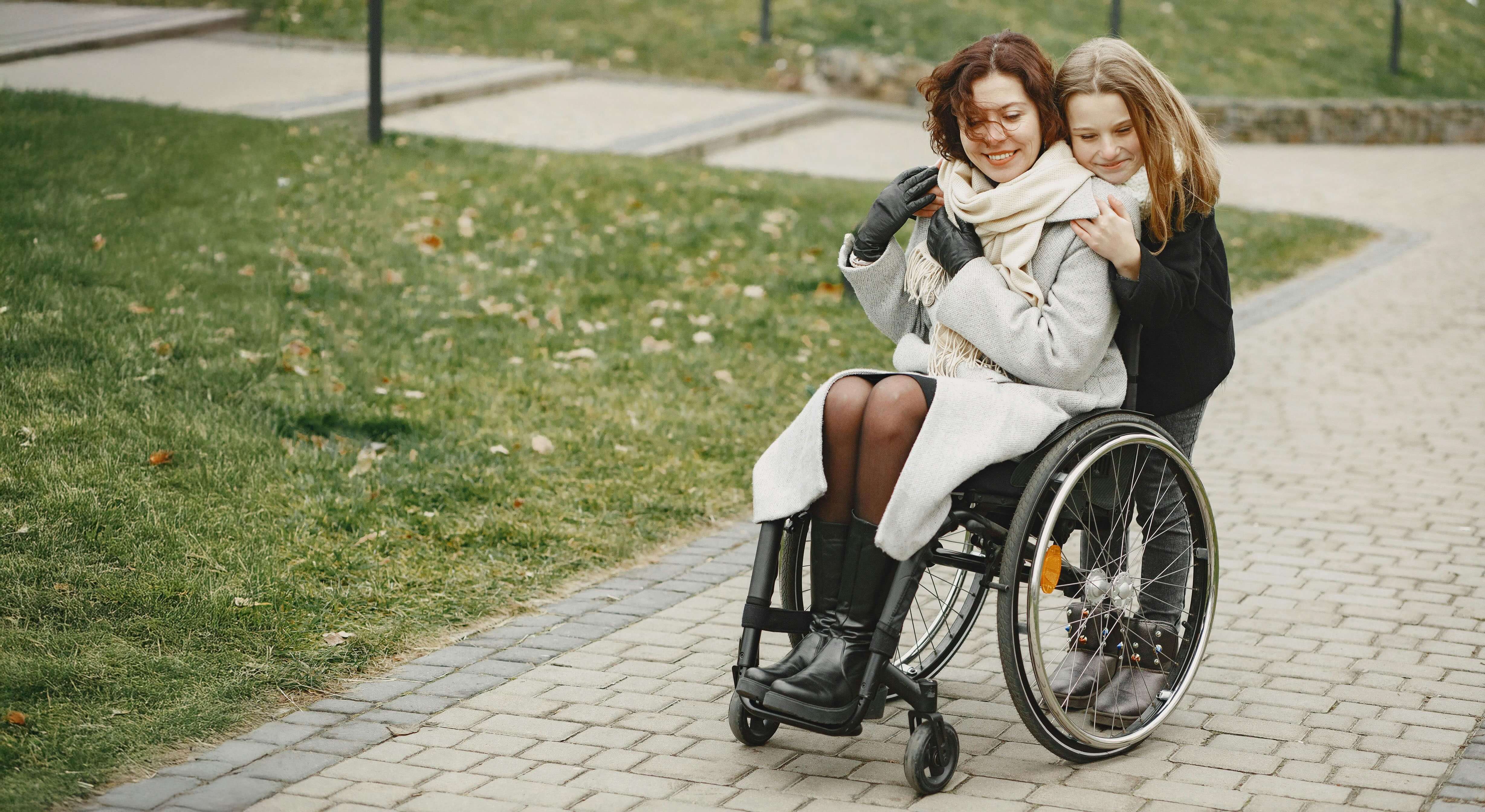 Two people, one in a wheelchair and the other pushing, on a paved path in a park.