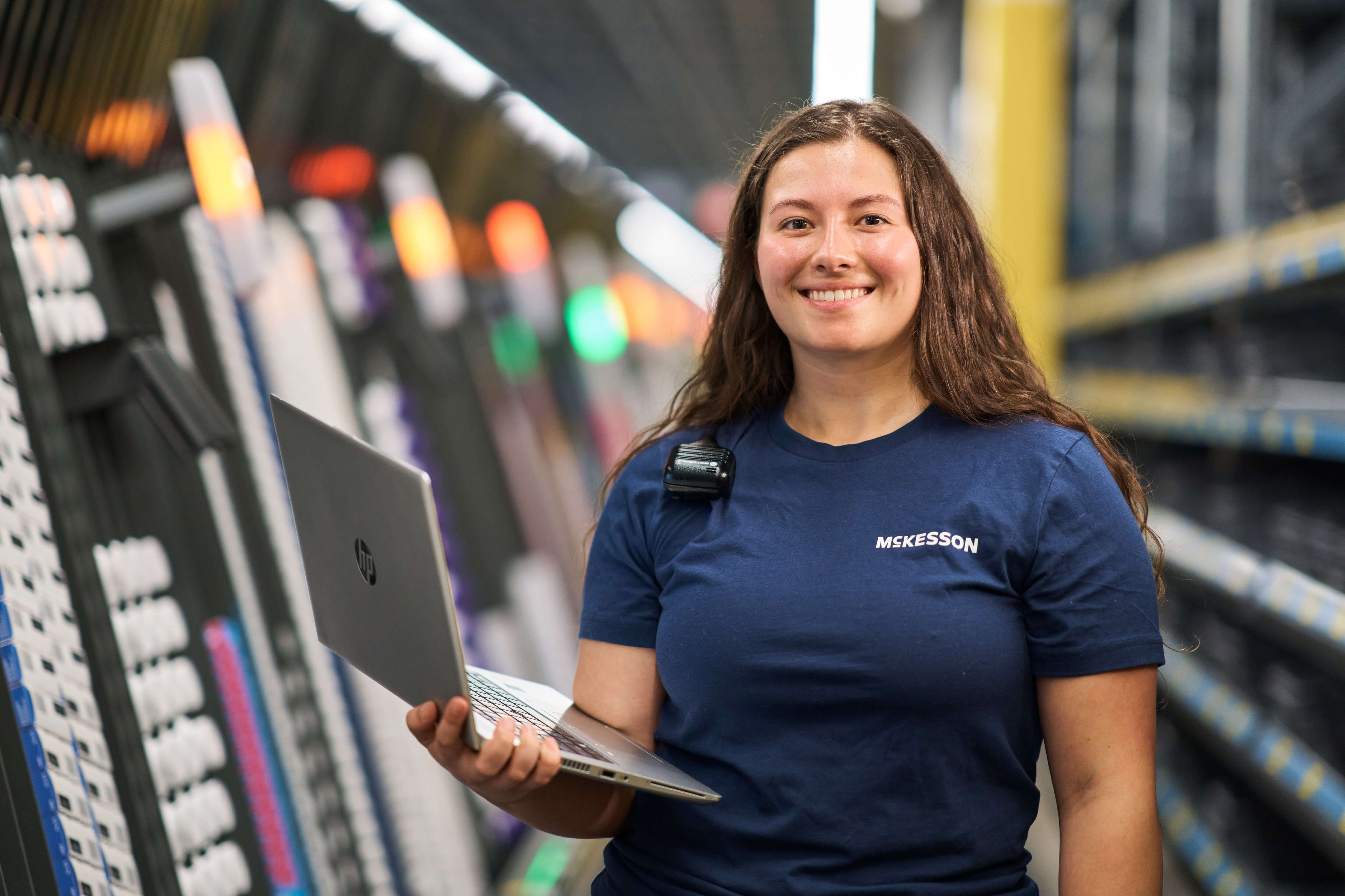 Person in a blue shirt holding a laptop, standing in an aisle with shelves on both sides.
