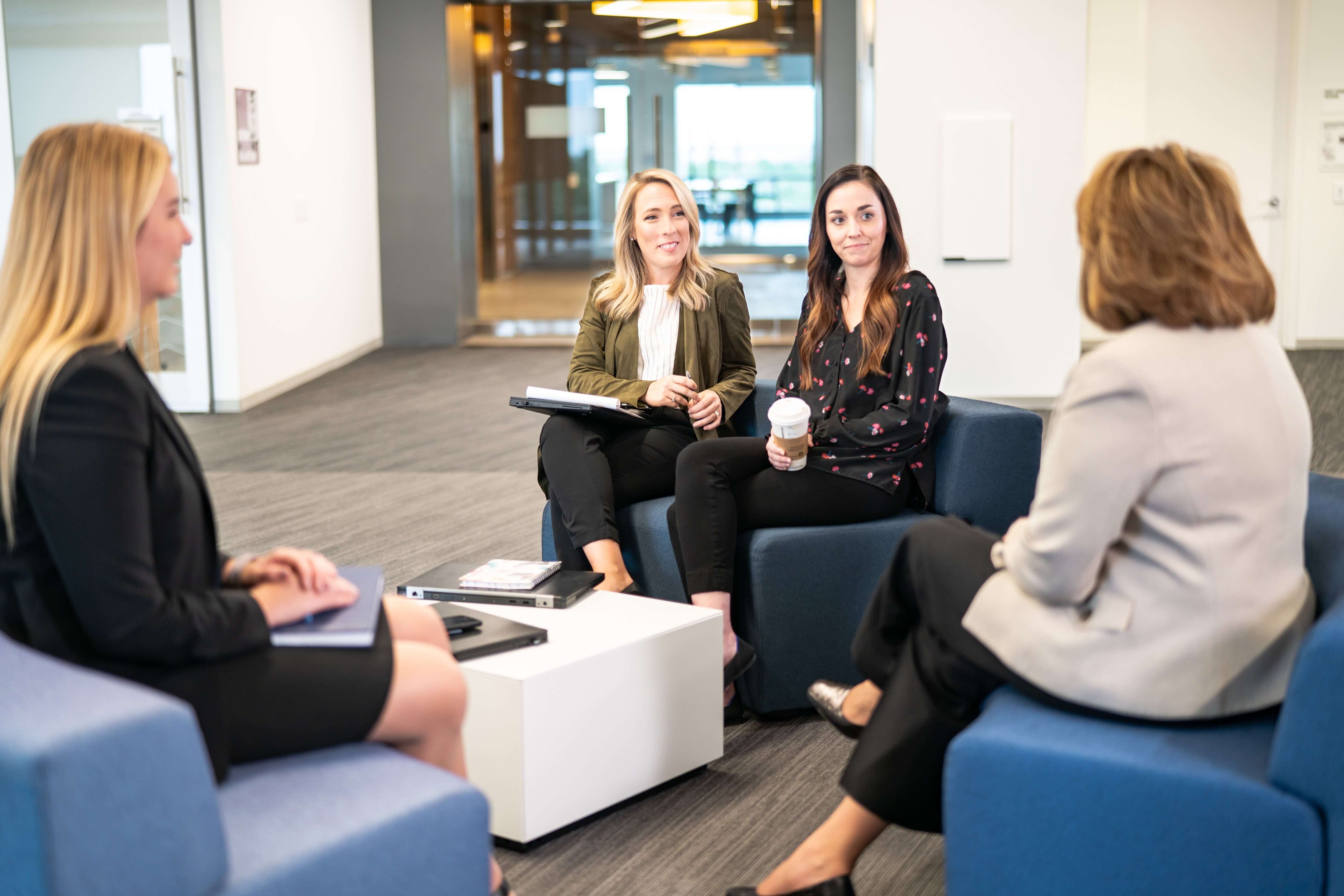 Four people in business attire are sitting and talking in a modern office lounge area.