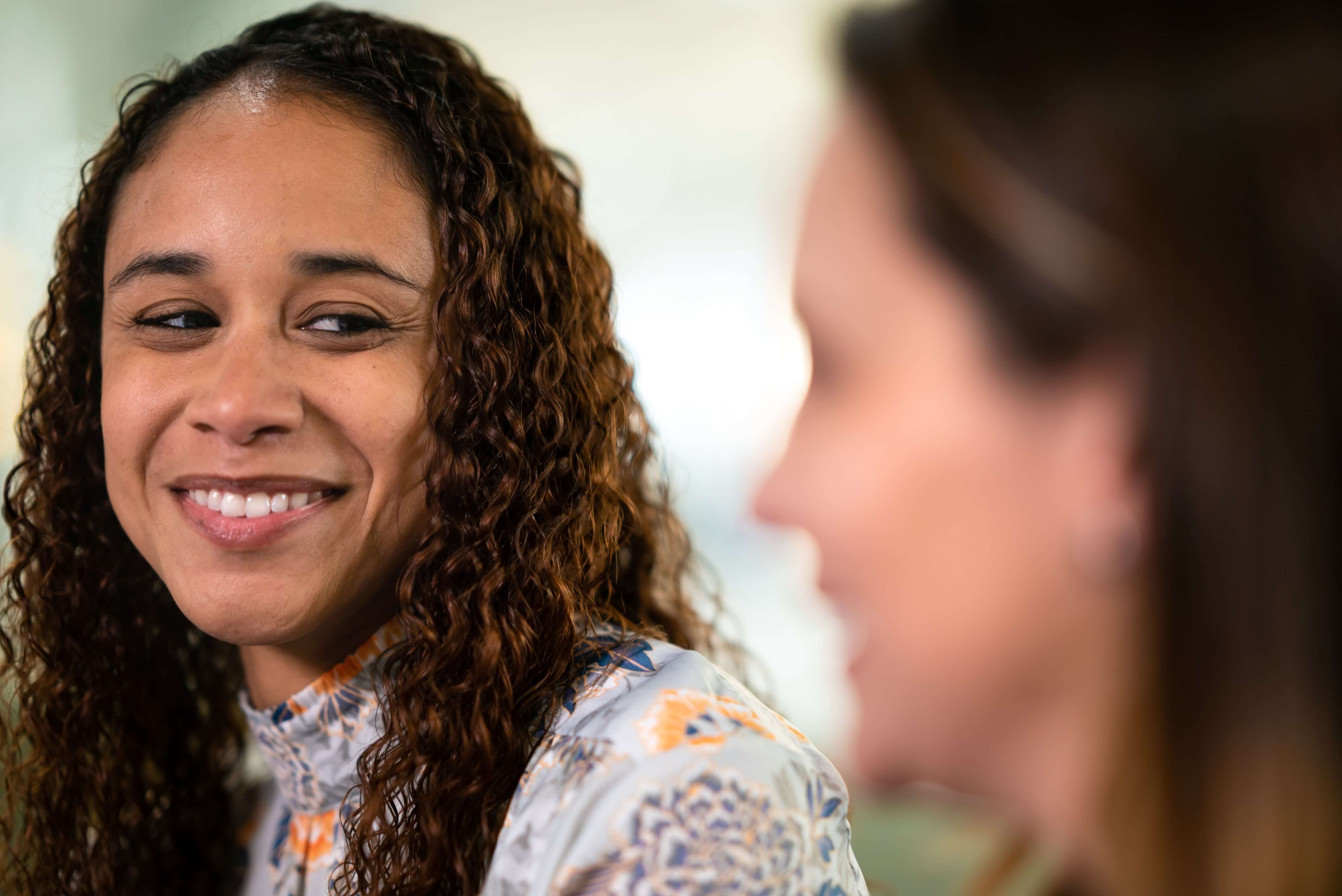Two people with blurred faces are talking, one with curly hair and a patterned shirt.