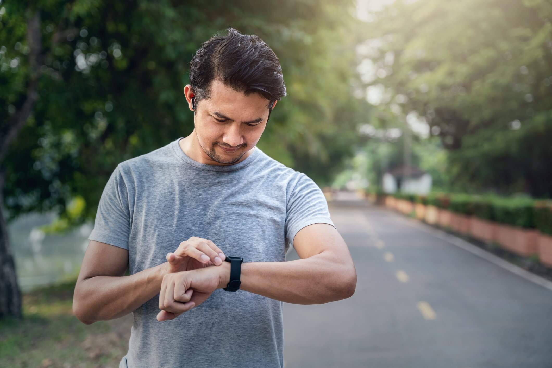A person in a gray shirt checks a smartwatch while standing on a tree-lined road.