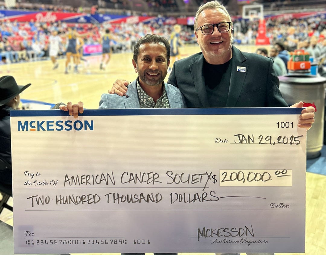 Two people hold a $200,000 McKesson check to the American Cancer Society at a basketball game with a crowd in the background.