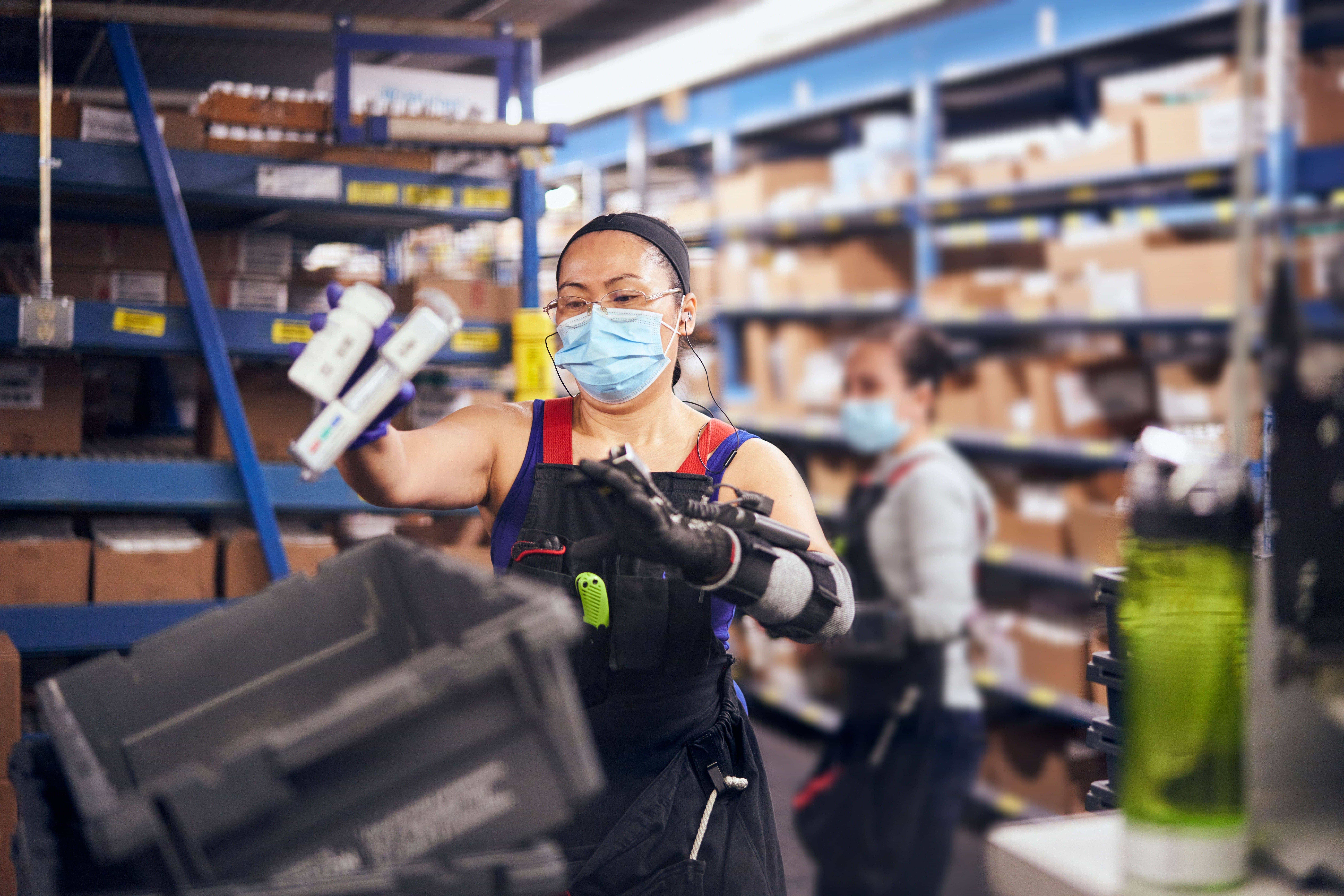 Two workers in a warehouse, one in the foreground placing items into a container, with shelves of boxes in the background.