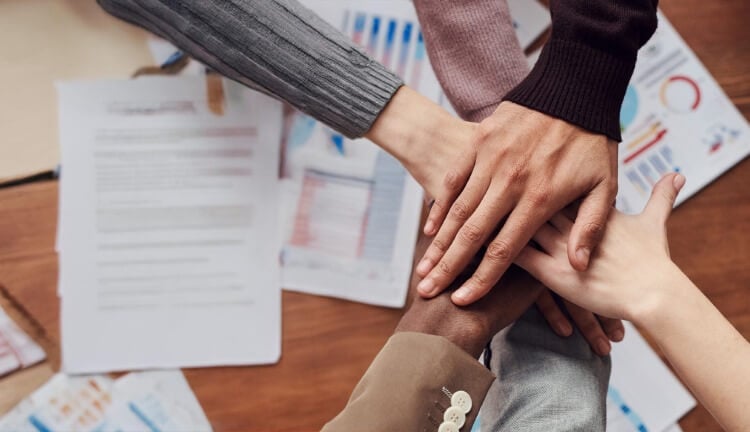 Five hands of diverse individuals placed on top of each other over a table with documents and charts.