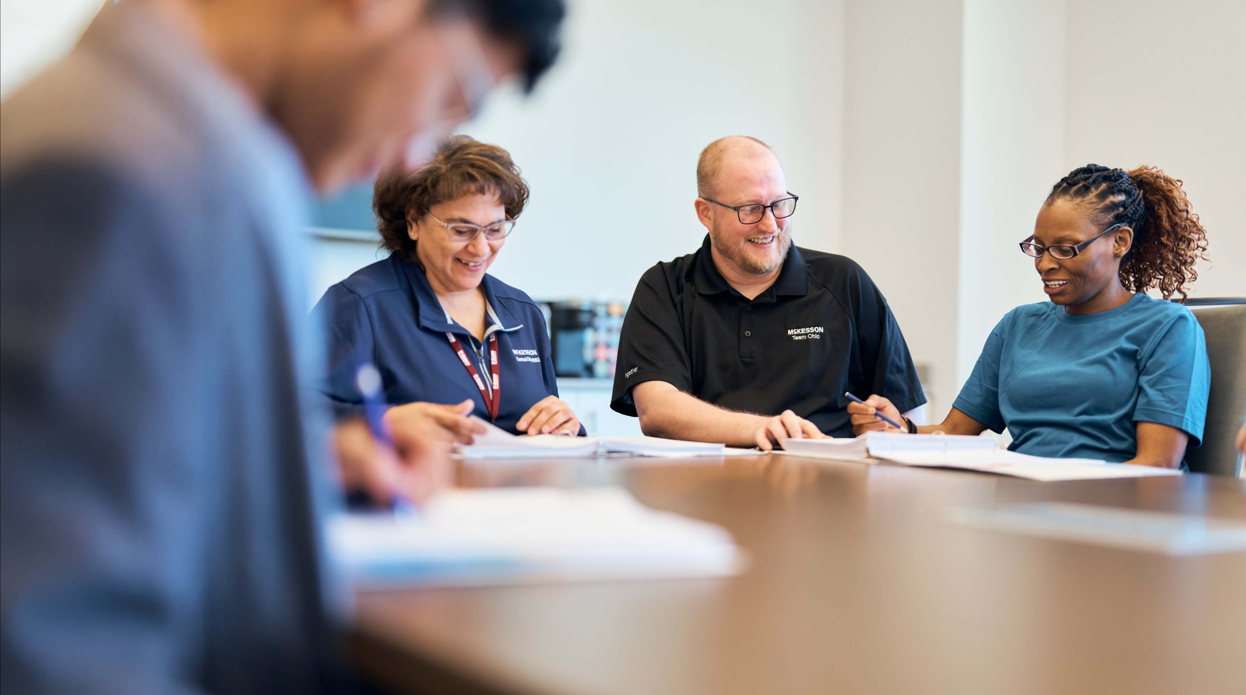 Four people sitting at a table in a meeting room, with one person in the foreground writing on paper.