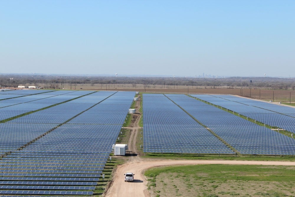 A large solar farm with rows of solar panels stretching into the distance, a dirt road, and a white vehicle in the foreground.