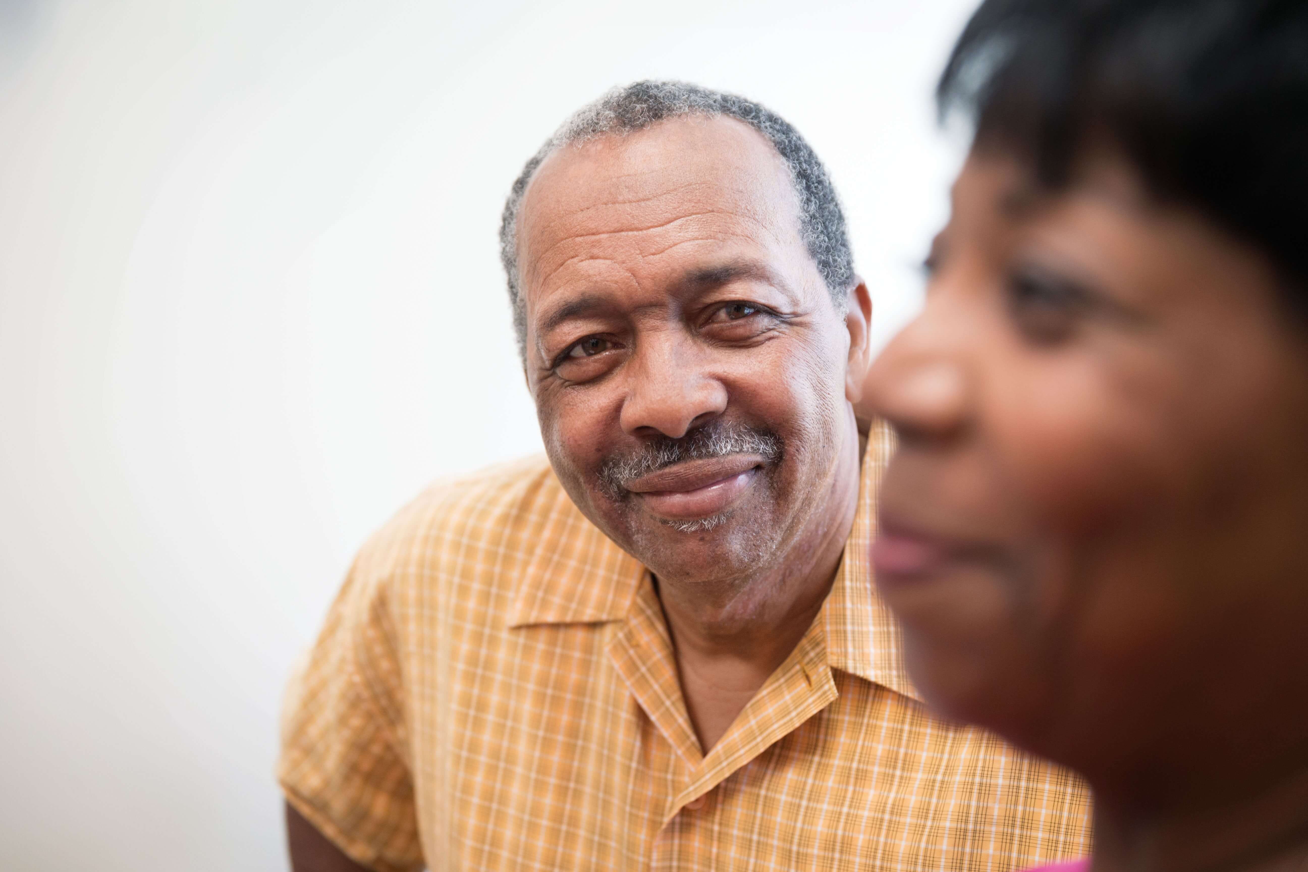 An older man smiling next to a woman against a plain wall.