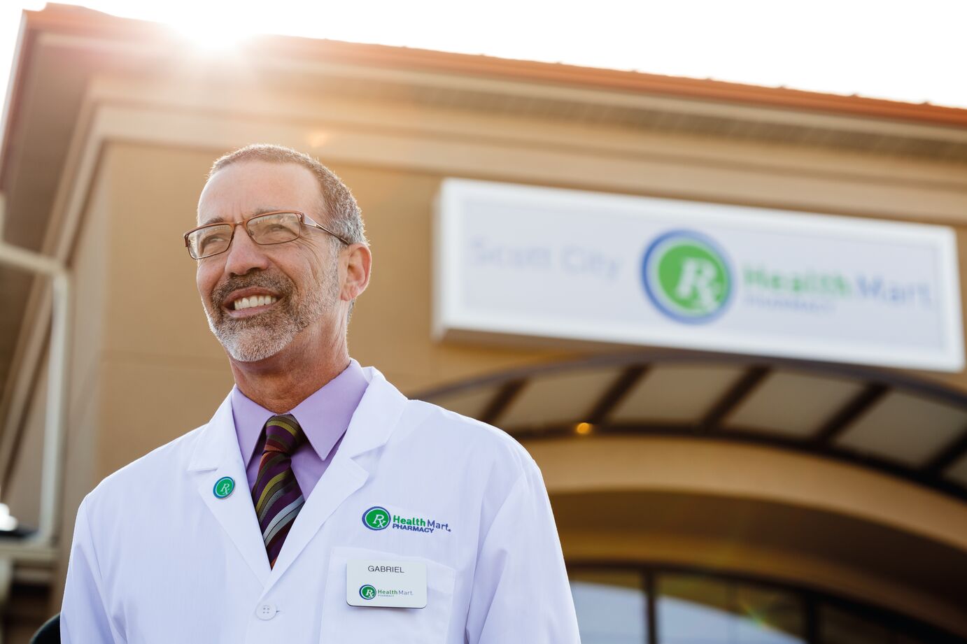 A person in a white lab coat stands in front of a building with a sign that reads 'Health Mart Pharmacy.'