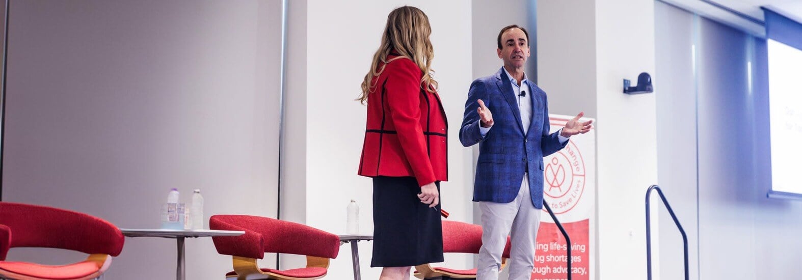 Two professionals on stage having a discussion, with red chairs and a banner about advocacy in the background.