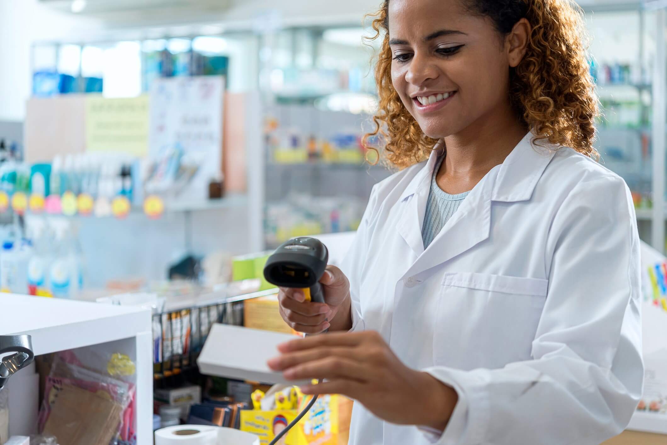 Pharmacist in a white coat scanning items at a pharmacy counter with shelves in the background.