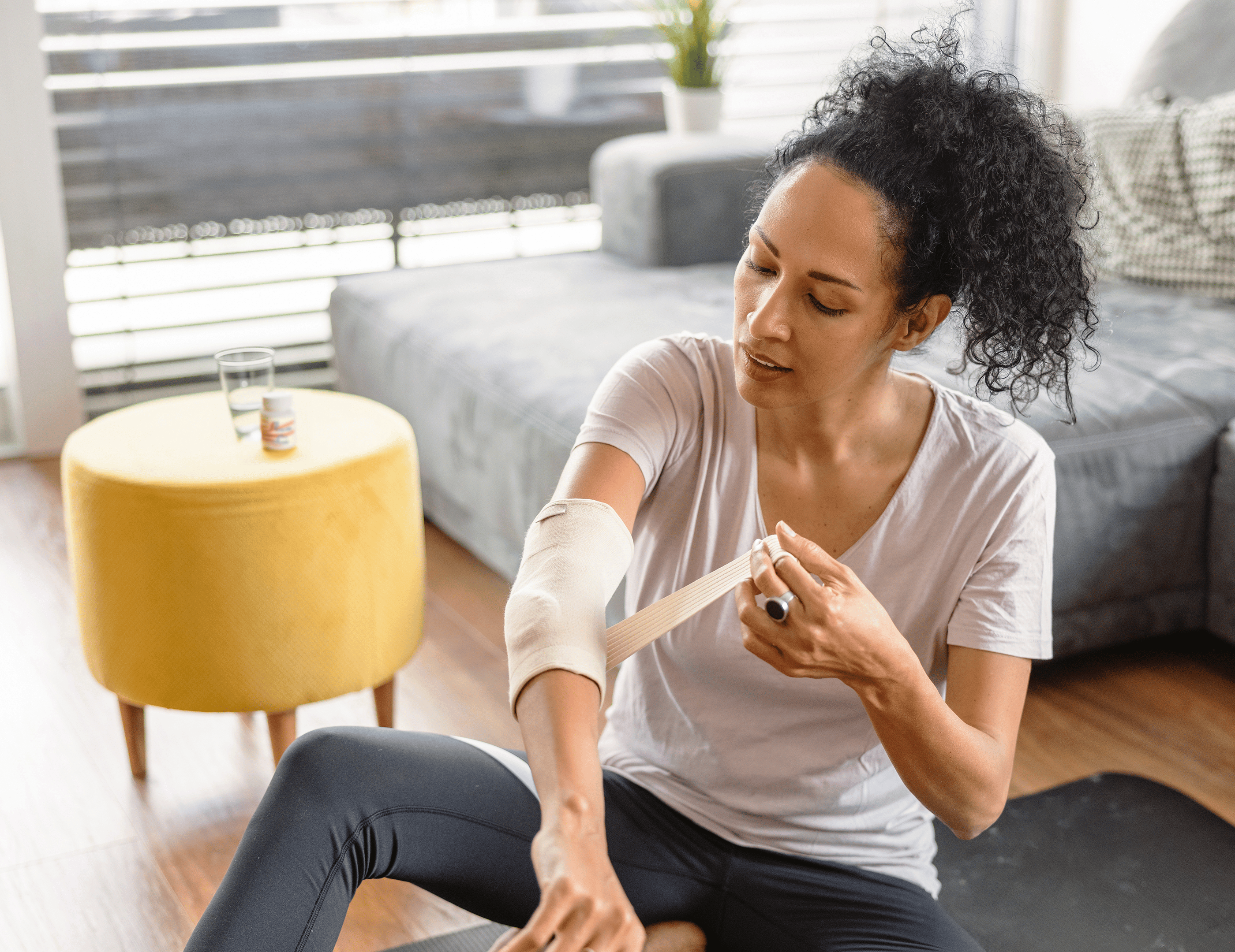 Person sitting on the floor in a living room, wrapping a bandage around their elbow. A yellow ottoman and a glass of water are in the background.