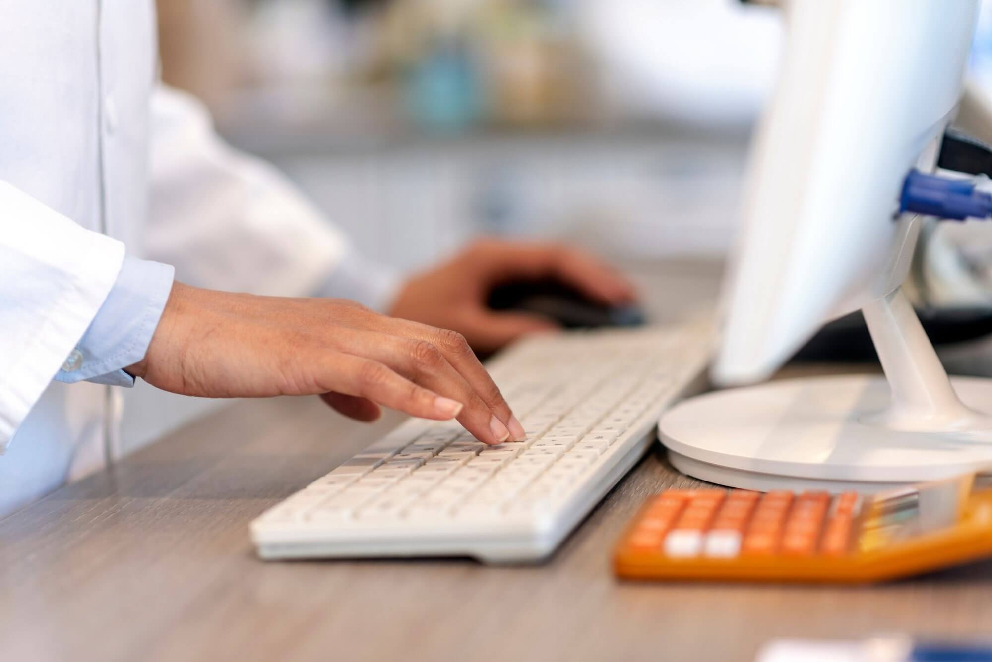 A person in a white coat typing on a keyboard with an orange calculator nearby.