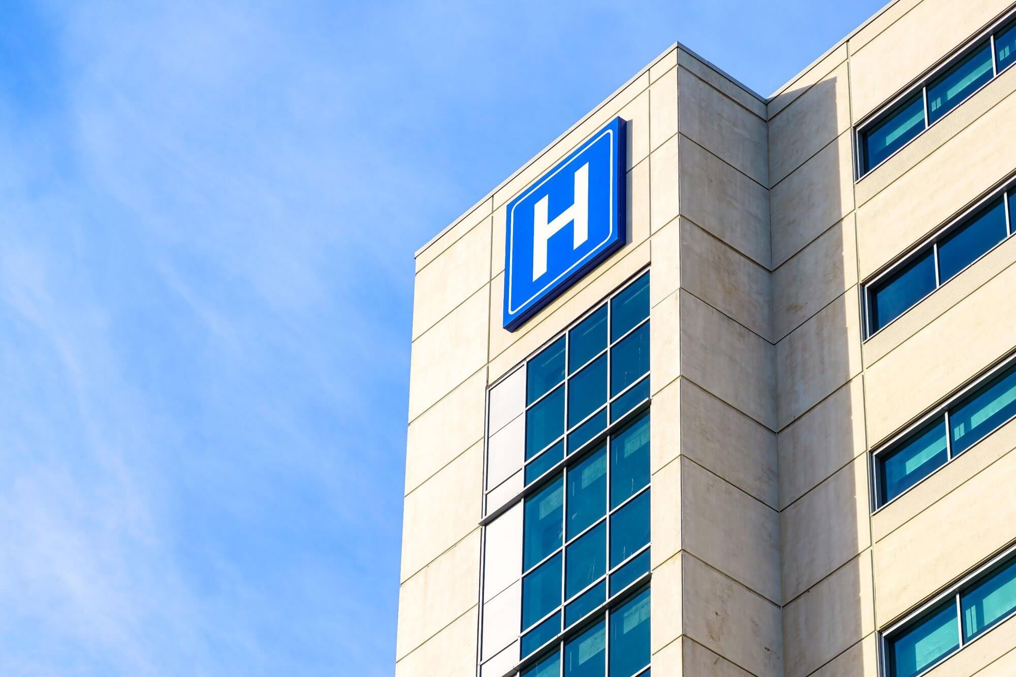 Hospital building with a blue 'H' sign on the upper corner against a clear sky.