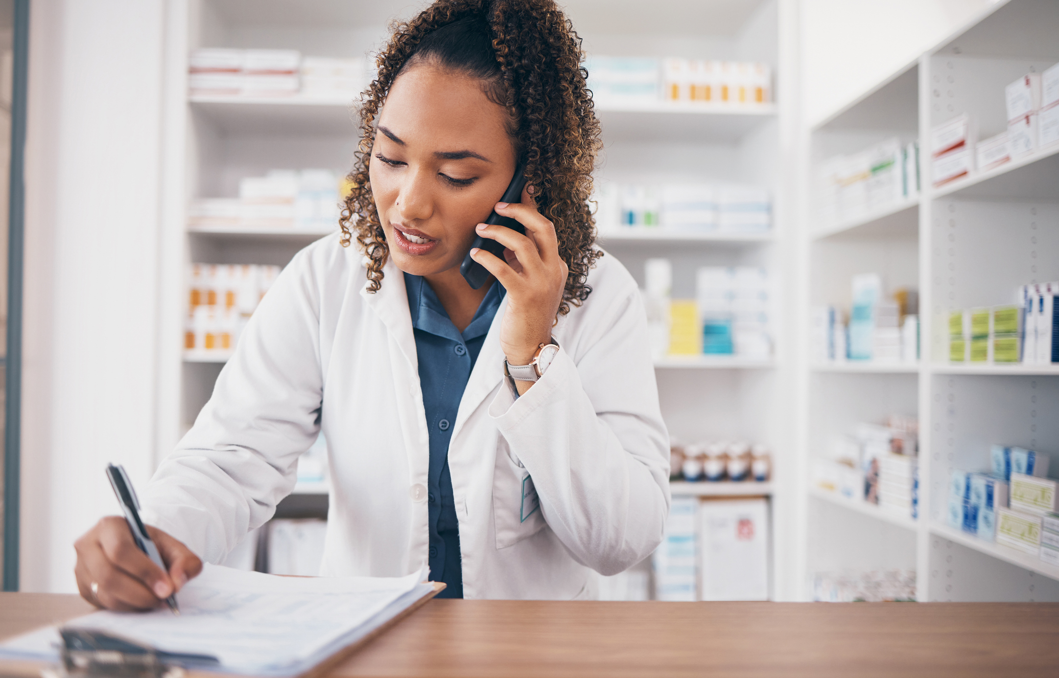 Pharmacist in a white coat on the phone, writing on a clipboard at a pharmacy counter with shelves of medications behind.