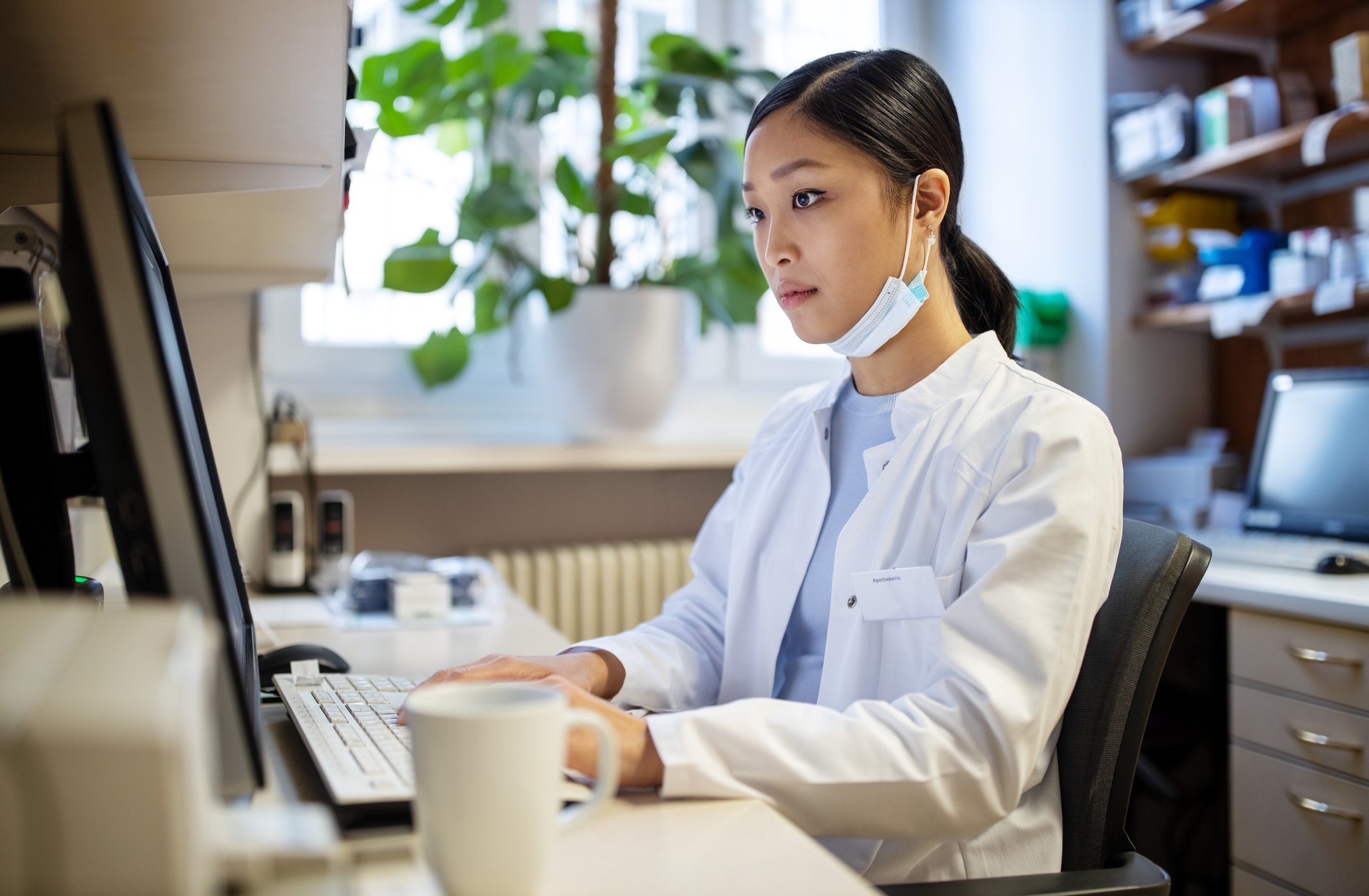 Person in a white lab coat working on a computer in an office with plants and shelves in the background.