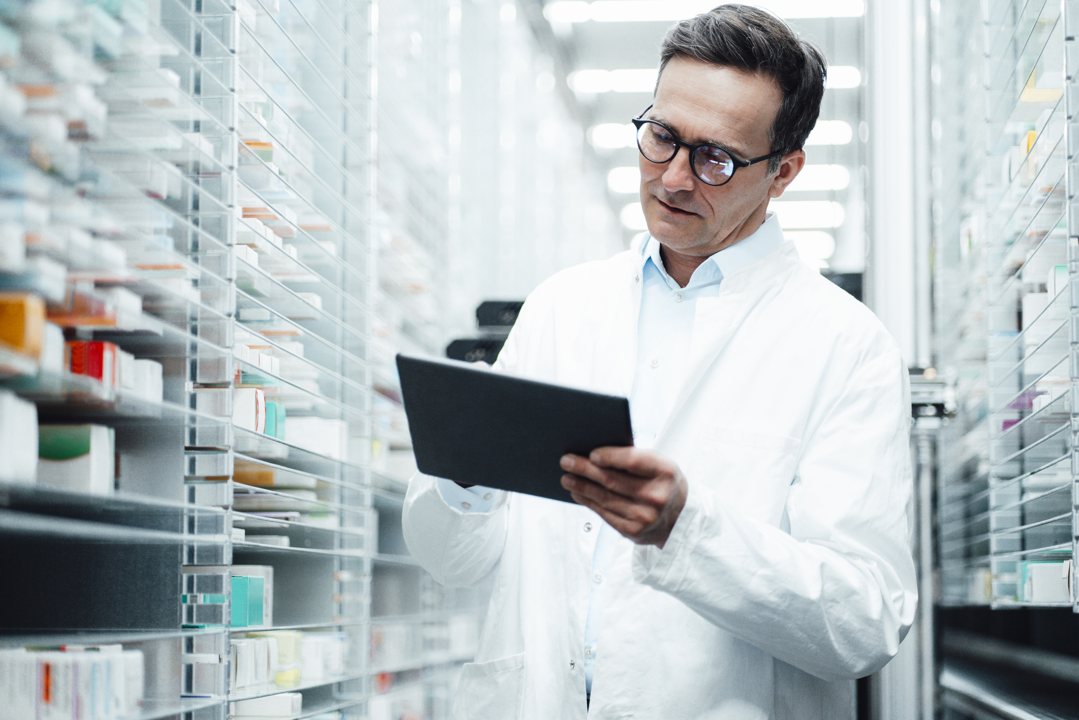 A person in a white lab coat holds a tablet while standing between shelves filled with various medical supplies in a well-lit storage area.