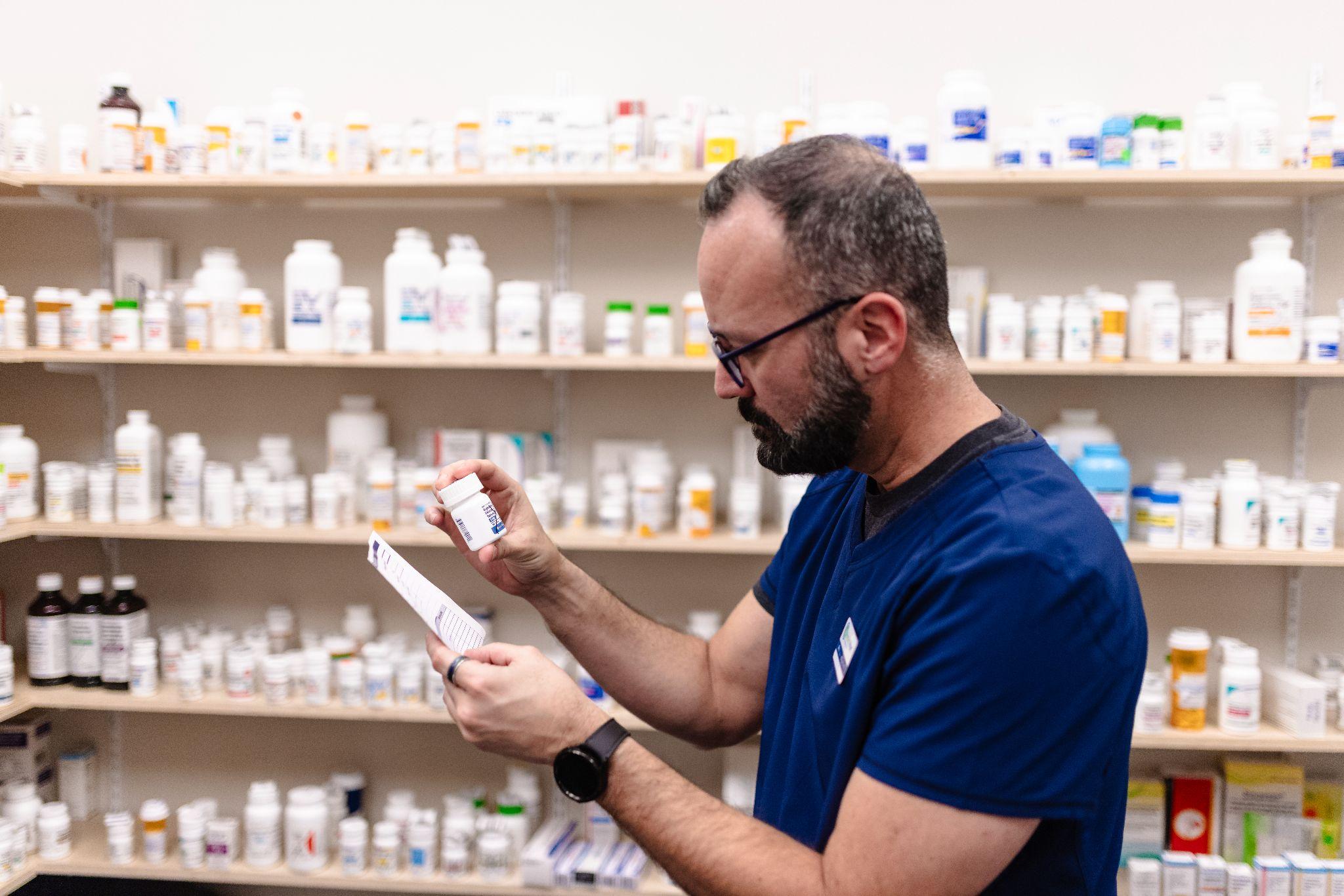 A person in a blue shirt holds a medication bottle and a paper, standing in front of shelves filled with various medication bottles in a pharmacy.