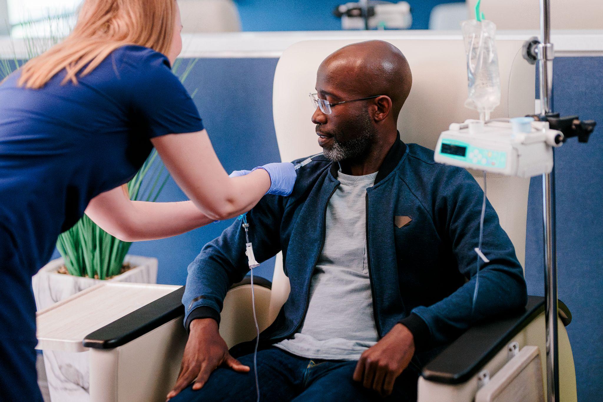 A healthcare professional administers an IV to a patient seated in a medical chair, with an IV drip and monitor nearby.