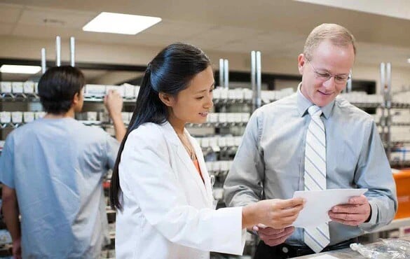 Pharmacists discussing documents in a medical supply room with shelves of medications in the background.