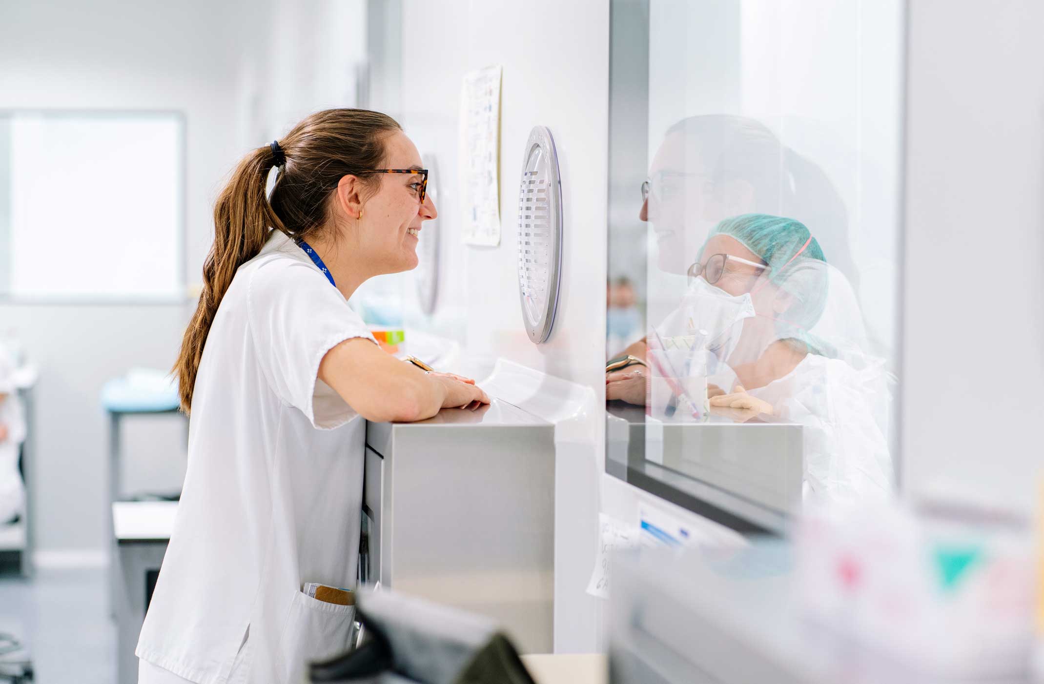 A healthcare worker in white scrubs leans on a counter, looking at a colleague through a glass partition.