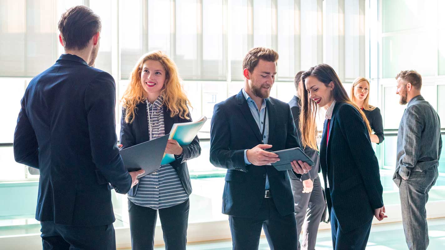 Group of business professionals in a bright office space, engaged in conversation and holding documents.