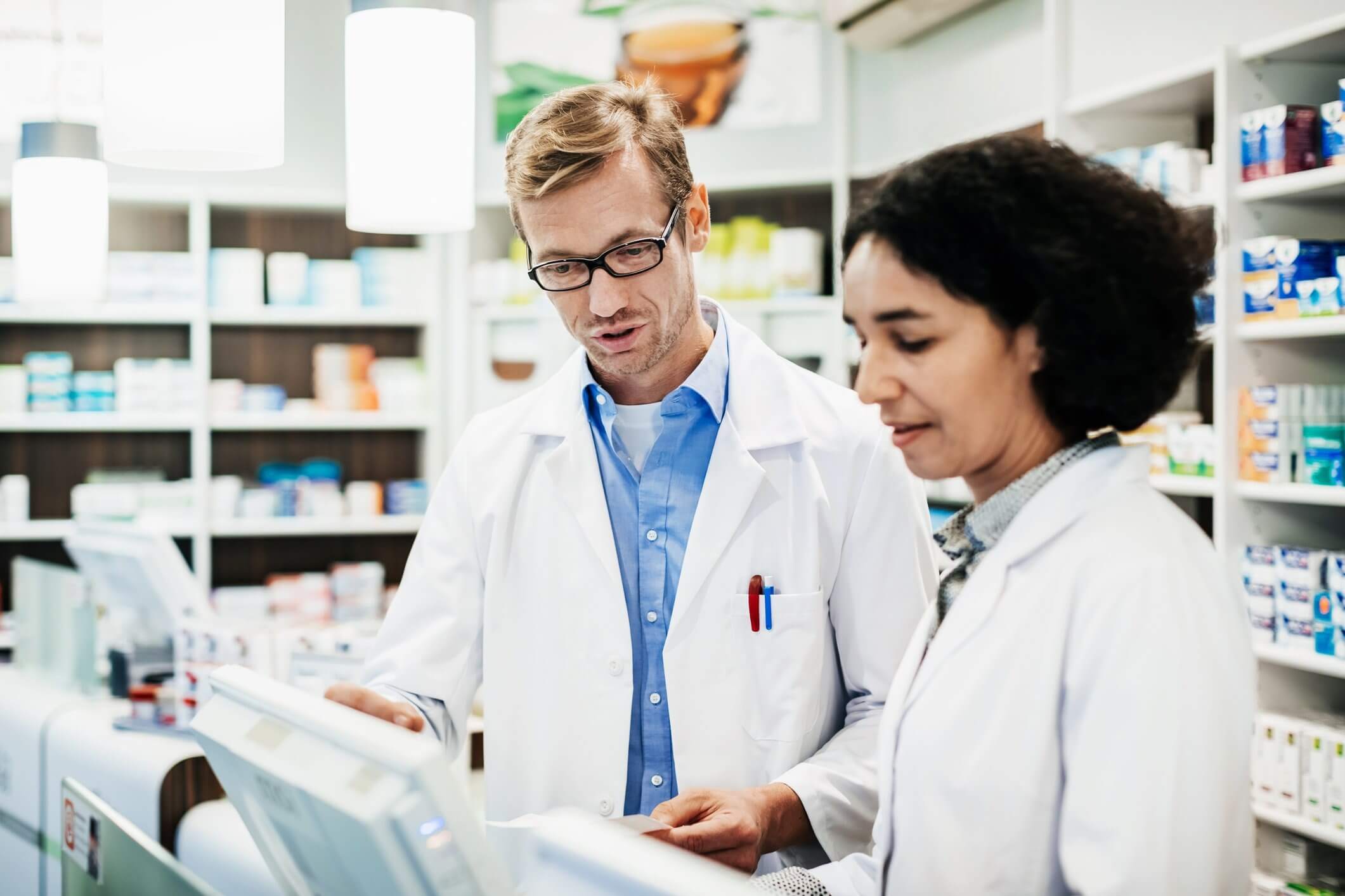 Two pharmacists in white lab coats stand behind a counter in a pharmacy, discussing something. Shelves with various medications are visible in the background
