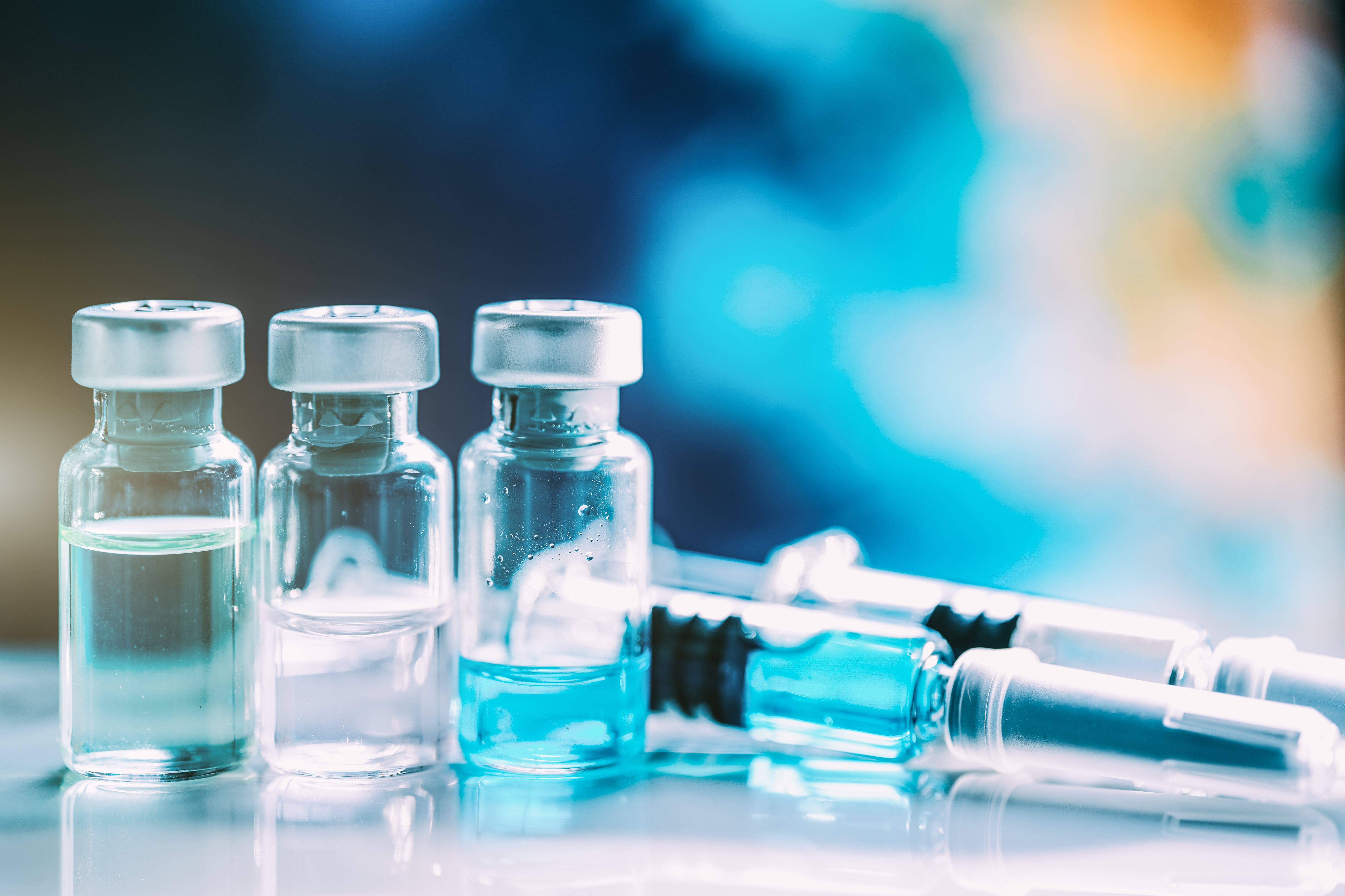 Three vials of liquid and two syringes on a reflective surface with a blurred blue and yellow background.