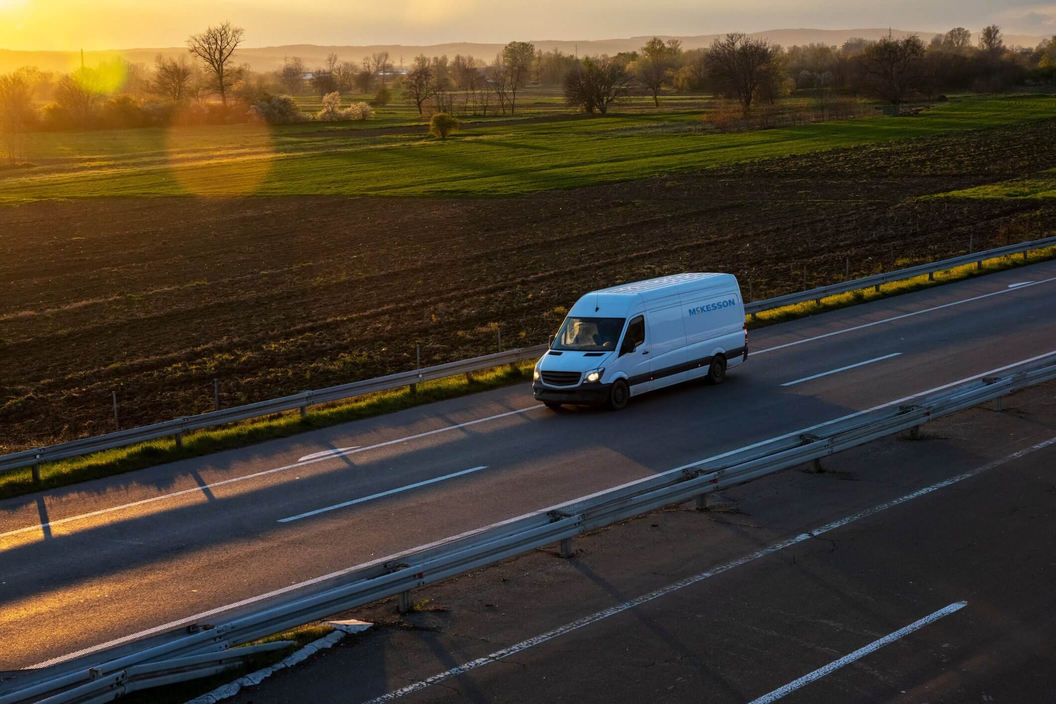 A white delivery van travels on a highway through a rural landscape at sunset, with green fields and bare trees in the background.