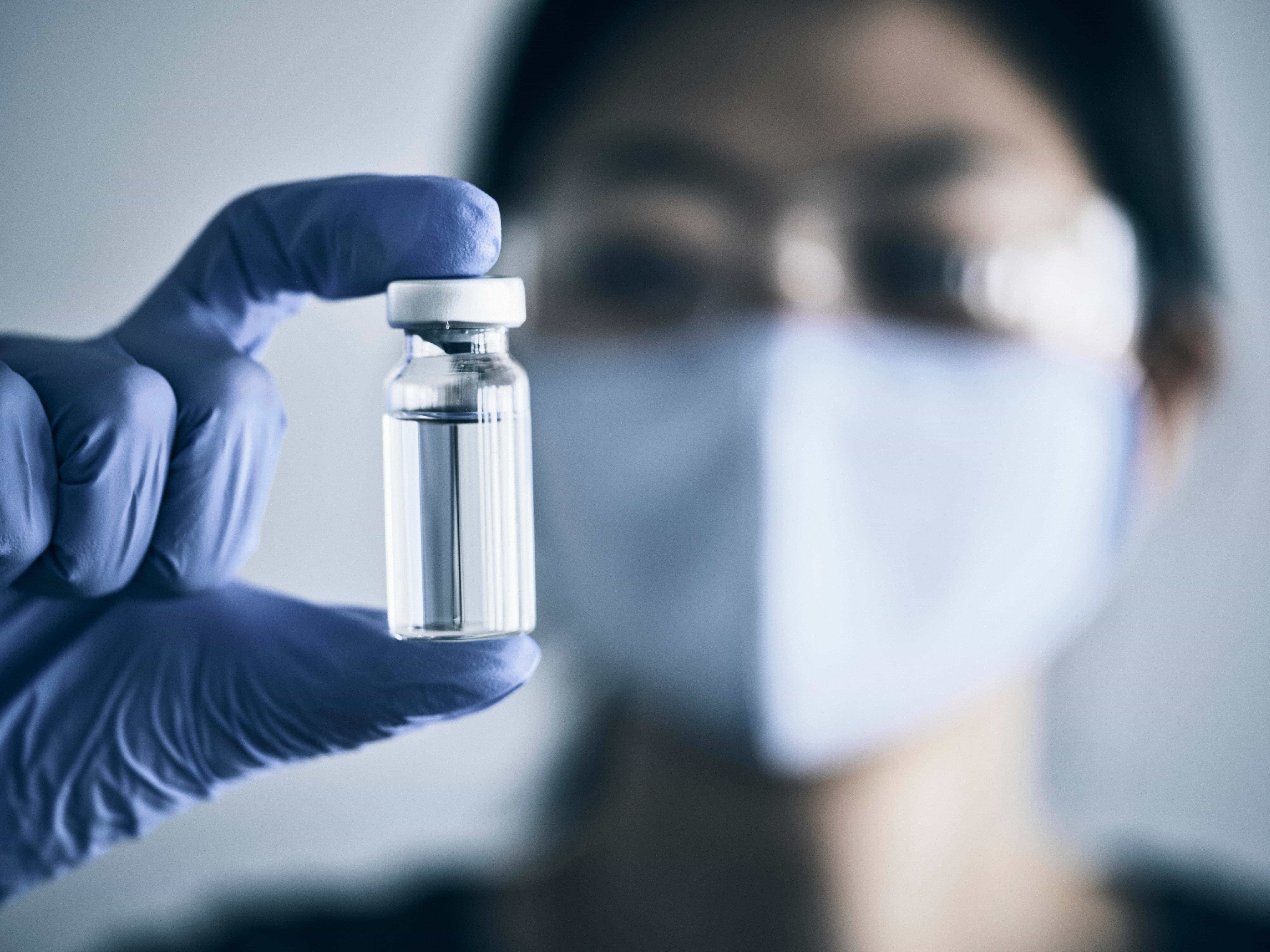 A gloved hand holds a small vial of clear liquid, with a blurred face in the background.