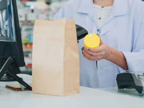 A person in a white lab coat scans a yellow bottle at a checkout counter with a brown paper bag nearby.