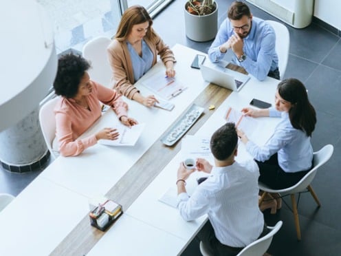 Five people having a meeting around a long table with laptops and documents.