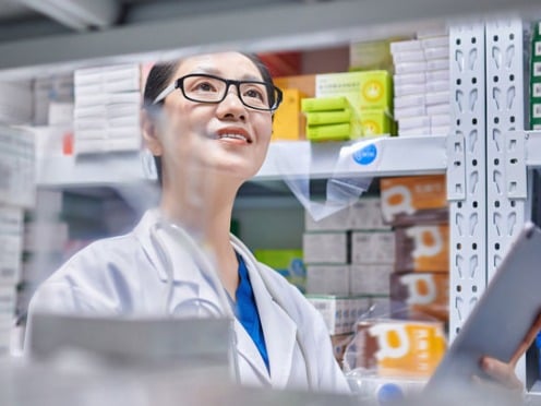 A pharmacist in a white coat holds a tablet while standing in front of shelves filled with medication boxes.