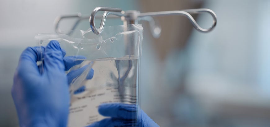 Gloved hands holding a clear IV bag with metal hooks visible in the background.