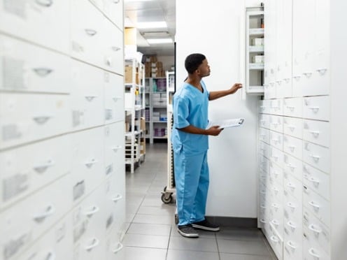 A healthcare professional in blue scrubs retrieves medication from a storage cabinet in a medical facility.