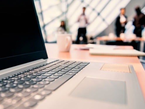 Close-up of a laptop on a desk in an office setting, with three blurred people in discussion in the background, conveying a professional atmosphere.
