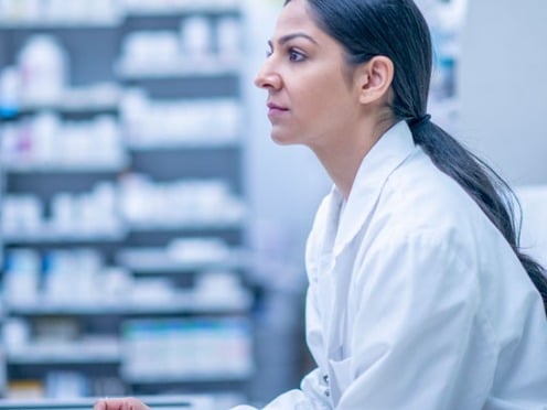 Person in a lab coat working at a computer in a pharmacy with shelves of medications in the background.