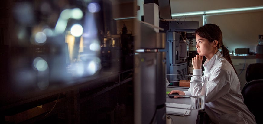 Person in a lab coat working at a computer in a dimly lit laboratory.